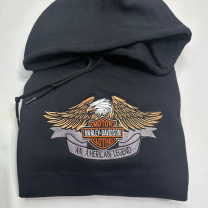A black pullover hoodie featuring a large embroidered Harley-Davidson winged logo patch on the front, set against a plain light background.