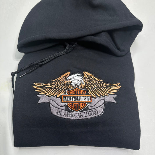 A black pullover hoodie featuring a large embroidered Harley-Davidson winged logo patch on the front, set against a plain light background.