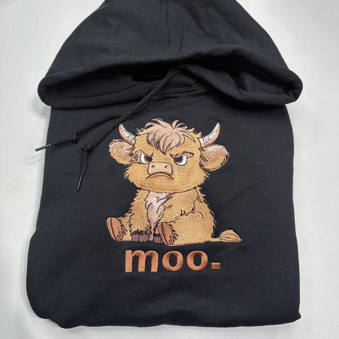 A black hoodie featuring an embroidered Scottish highland cow character with the text 'moo-' embroidered below it, displayed on a light gray background.