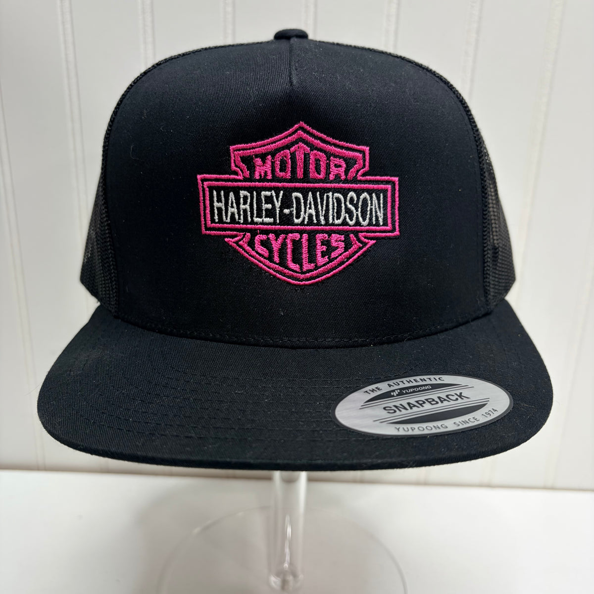 Black snapback trucker hat with a pink embroidered Harley-Davidson logo patch on the front, displayed on a clear stand against a white paneled background.