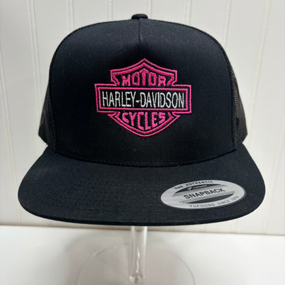 Black snapback trucker hat with a pink embroidered Harley-Davidson logo patch on the front, displayed on a clear stand against a white paneled background.