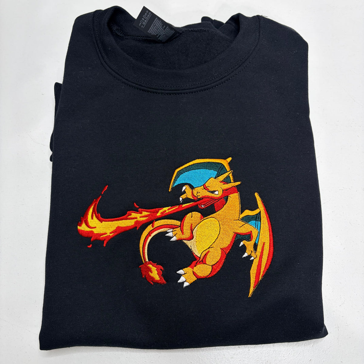 A folded black crewneck sweatshirt featuring a colorful embroidered graphic of Charizard on the front, set against a plain white background.
