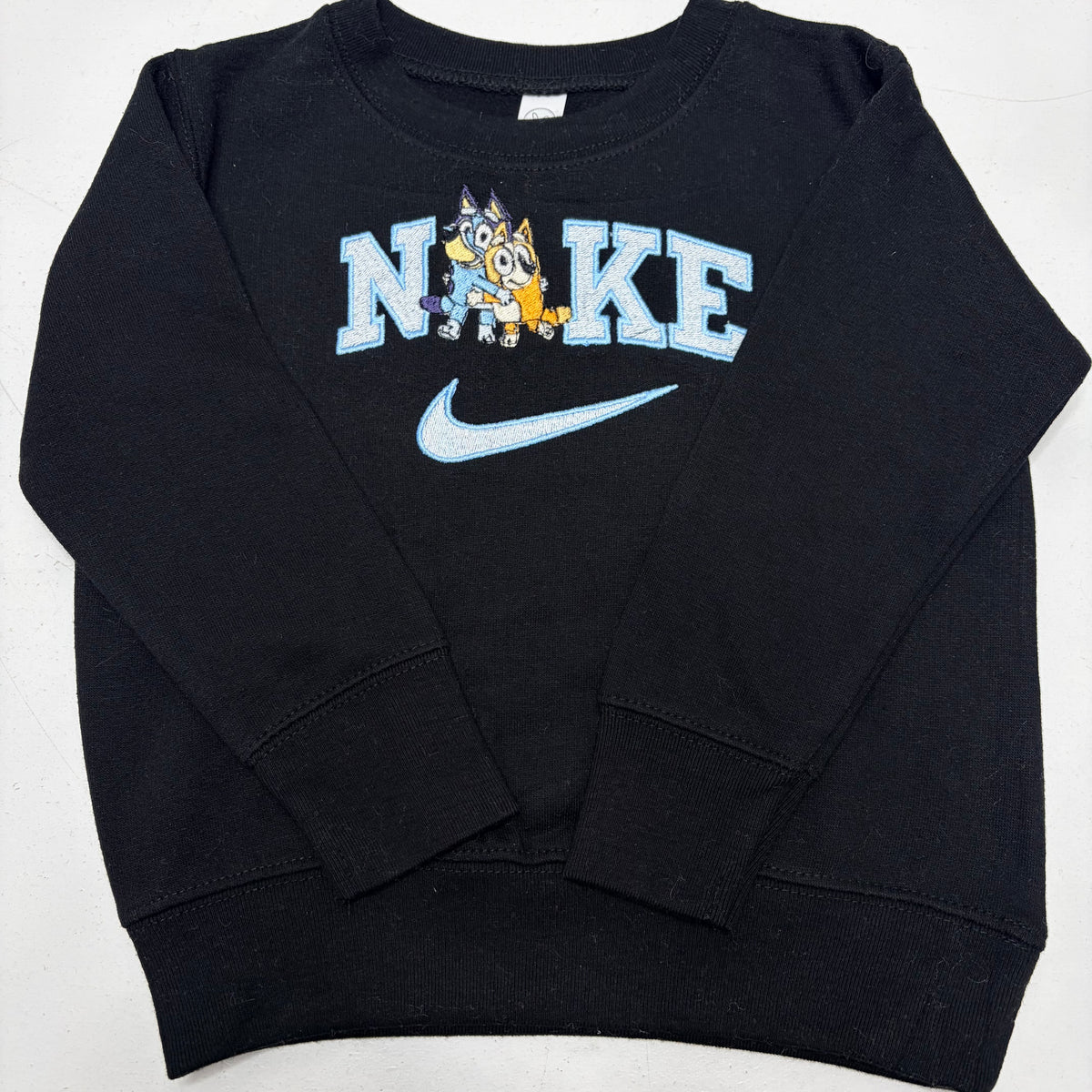A black long-sleeve sweatshirt featuring a light blue 'NIKE' graphic with a cartoon bluey dog mascot and a light blue swoosh on the front.