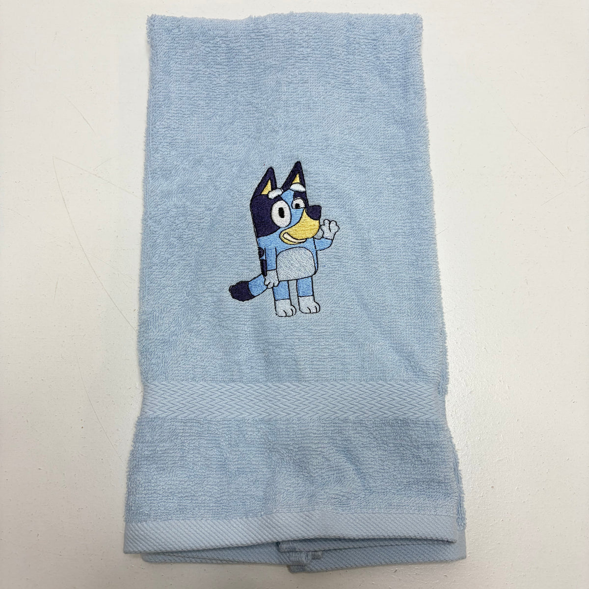 A light blue terry cloth towel featuring an embroidered Bluey character on the front, displayed flat against a plain off-white background.