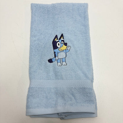 A light blue terry cloth towel featuring an embroidered Bluey character on the front, displayed flat against a plain off-white background.