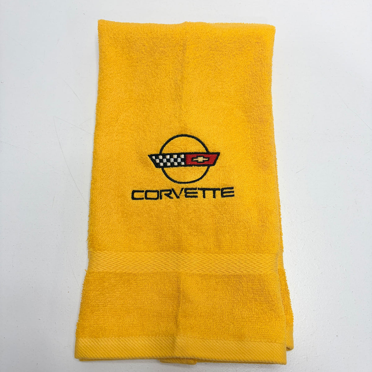 A folded yellow terry cloth towel featuring an embroidered Corvette logo and text in the center, set against a plain white background.