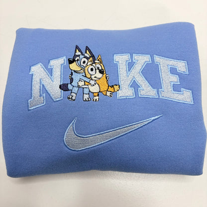A folded light blue sweatshirt featuring an embroidered design with the word 'NiKE' in light blue varsity letters, a cartoon bluey and bingo dog characters in the center, and a gray embroidered Nike swoosh below.