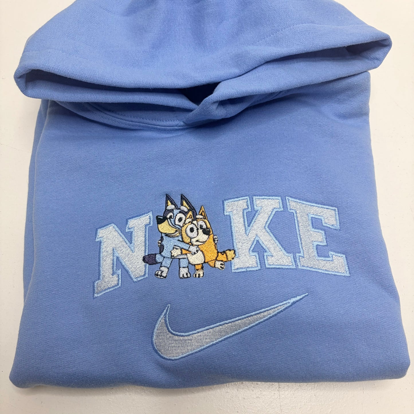 A folded light blue Nike hoodie featuring a custom embroidered design with the word 'NIKE' stylized to include a cartoon bluey and bingo dog characters, and a silver-gray swoosh below.