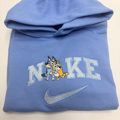 A folded light blue Nike hoodie featuring a custom embroidered design with the word 'NIKE' stylized to include a cartoon bluey and bingo dog characters, and a silver-gray swoosh below.