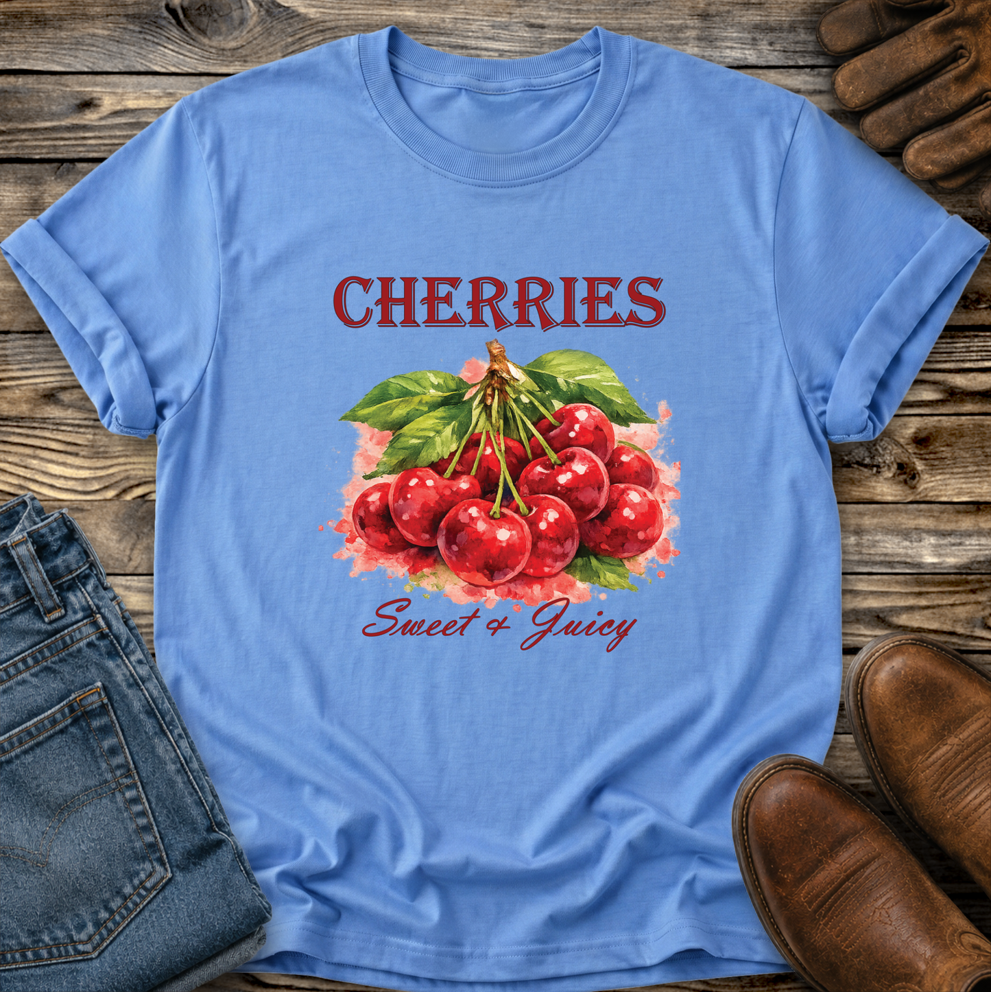 A light blue graphic t-shirt featuring a watercolor illustration of red cherries with green leaves and the text 'CHERRIES Sweet & Juicy'. The t-shirt is styled in a flat-lay arrangement on a rustic wooden background, accompanied by blue jeans, brown leather shoes, and leather gloves.