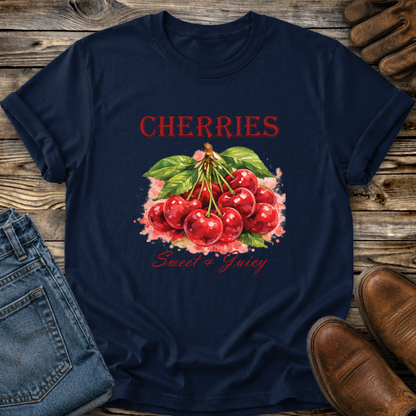 A navy blue graphic t-shirt featuring a watercolor illustration of red cherries with green leaves and pink blossoms, with the text 'CHERRIES Sweet & Juicy' printed on the front. The t-shirt is styled in a flat-lay arrangement on a rustic wooden background, accompanied by blue jeans, brown leather shoes, and leather gloves.