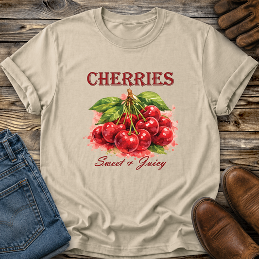 A beige graphic t-shirt featuring a watercolor illustration of red cherries with green leaves and the text 'CHERRIES Sweet & Juicy'. The t-shirt is styled in a flat-lay arrangement on a rustic wooden background, accompanied by blue jeans, brown leather shoes, and a leather glove.