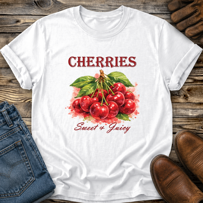 A white short-sleeve T-shirt laid flat on a rustic wooden surface, featuring a red cherry graphic with green leaves and the words 'CHERRIES' and 'Sweet & Juicy' in red script. Blue jeans, brown shoes, and a small brown accessory are placed around the shirt as styling props.