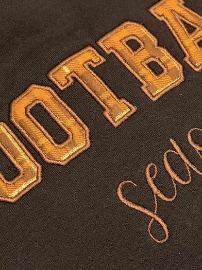 Close-up of a dark brown or black fabric with large metallic gold embroidered lettering and smaller script embroidery.