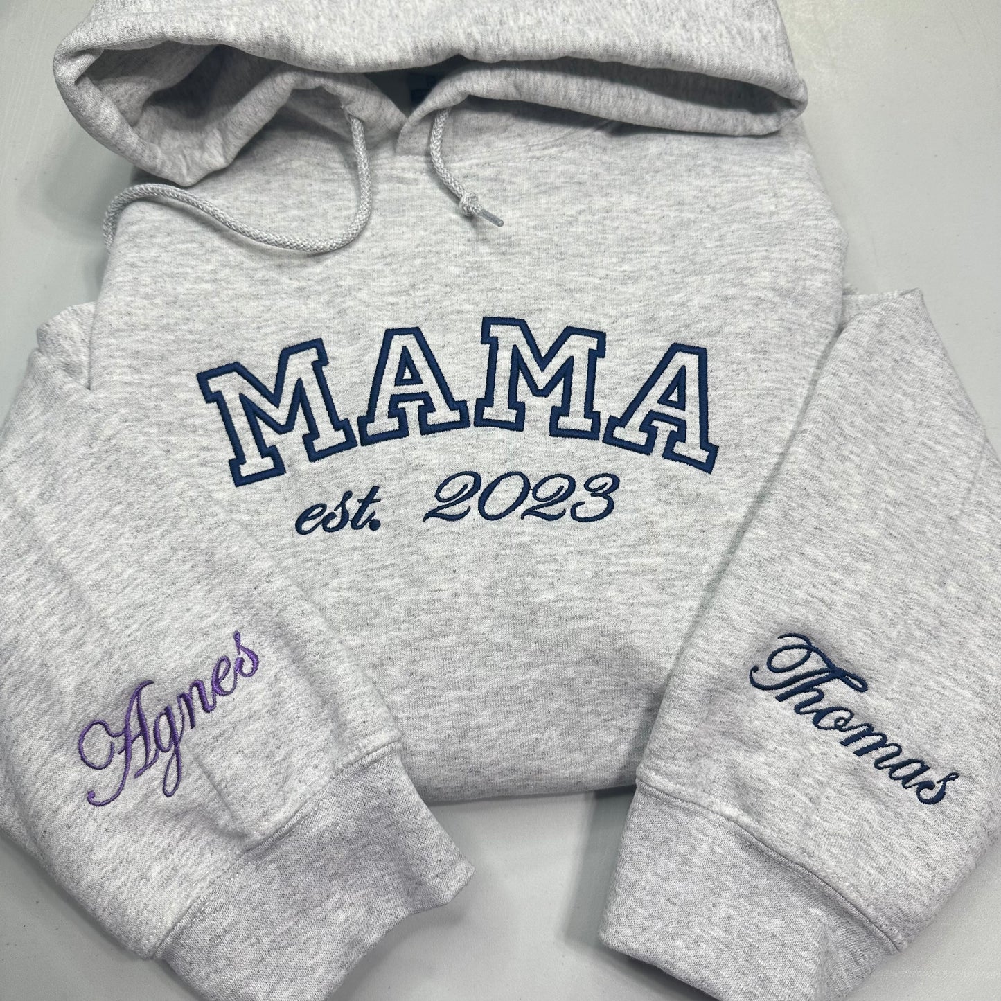 A heather gray hoodie laid flat on a light gray background, with large navy outlined 'MAMA' text across the chest and smaller 'est. 2023' below it.