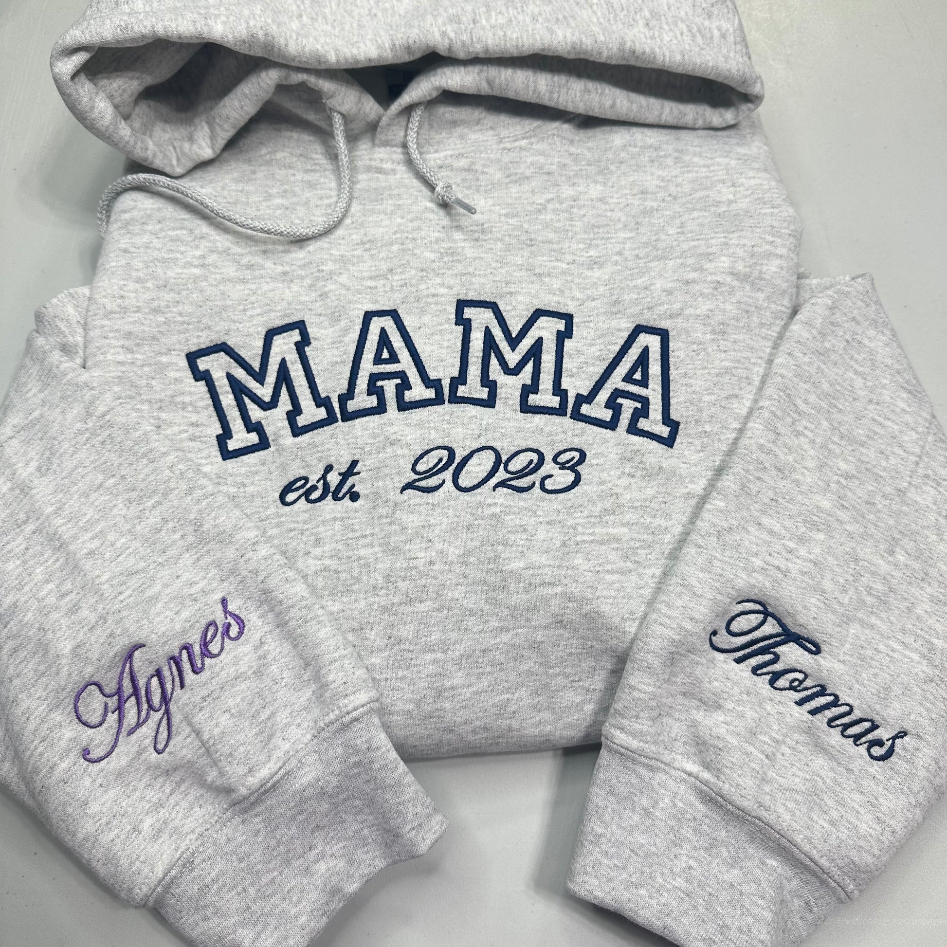 A heather gray hoodie laid flat on a light gray background, with large navy outlined 'MAMA' text across the chest and smaller 'est. 2023' below it.
