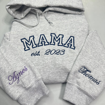 A heather gray hoodie laid flat on a light gray background, with large navy outlined 'MAMA' text across the chest and smaller 'est. 2023' below it.