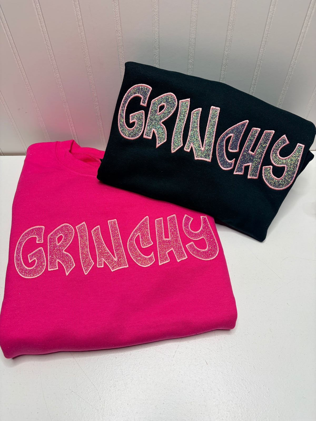 Two folded sweatshirts with the word 'GRINCHY' in large glittery letters, one bright pink and one black, displayed on a white surface against a white paneled wall.