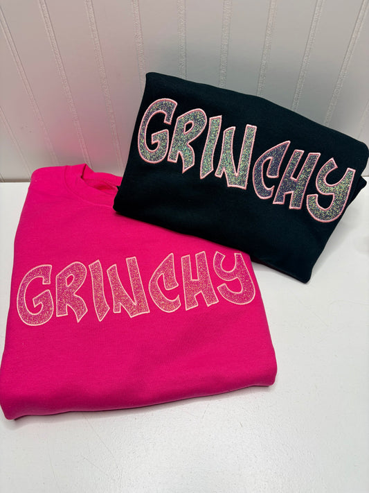 Two folded sweatshirts with the word 'GRINCHY' in large glittery letters, one bright pink and one black, displayed on a white surface against a white paneled wall.