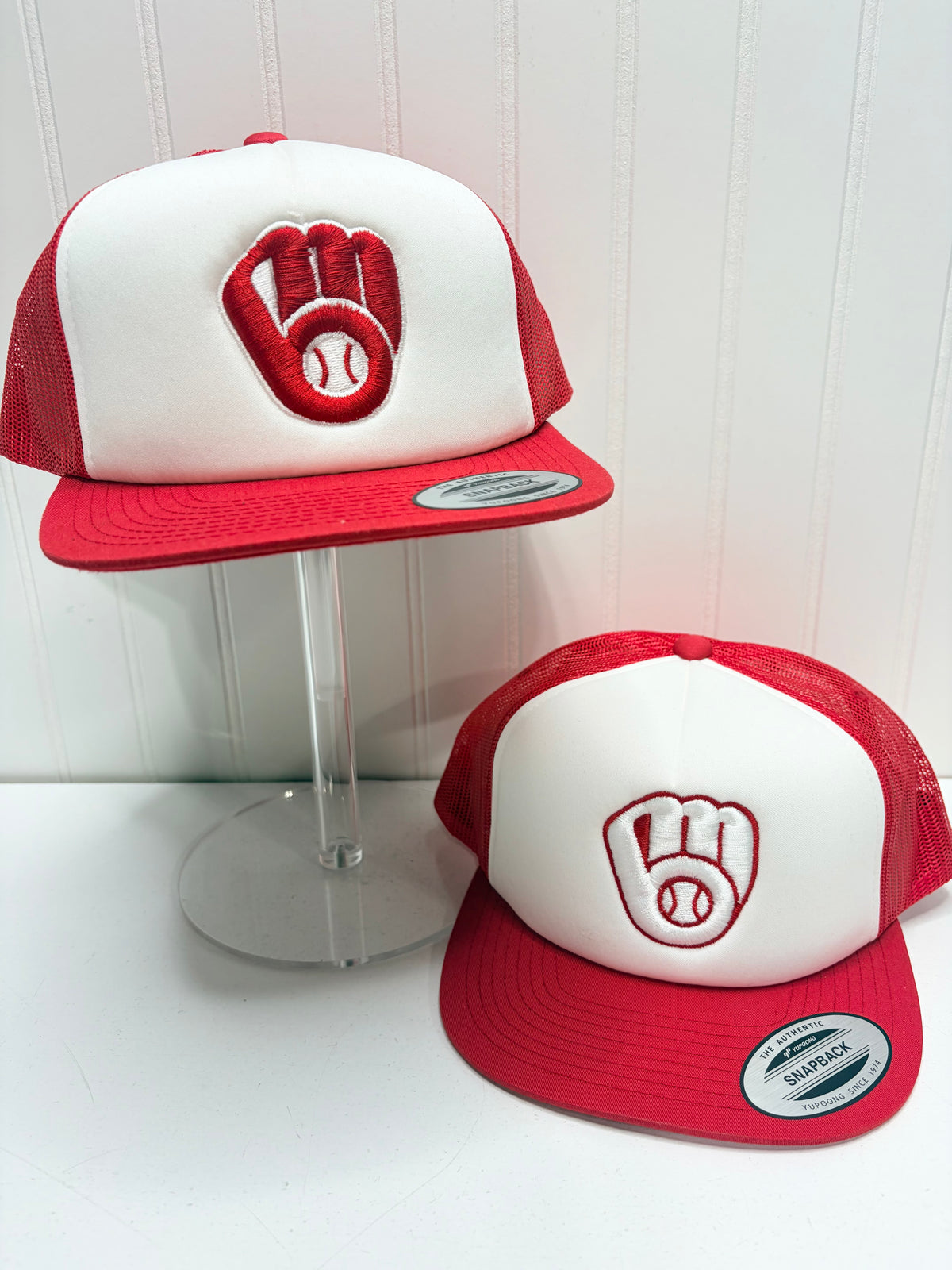 Two red and white trucker caps with a baseball glove and baseball logo on the front, displayed on a clear stand and a white surface against a white paneled wall.