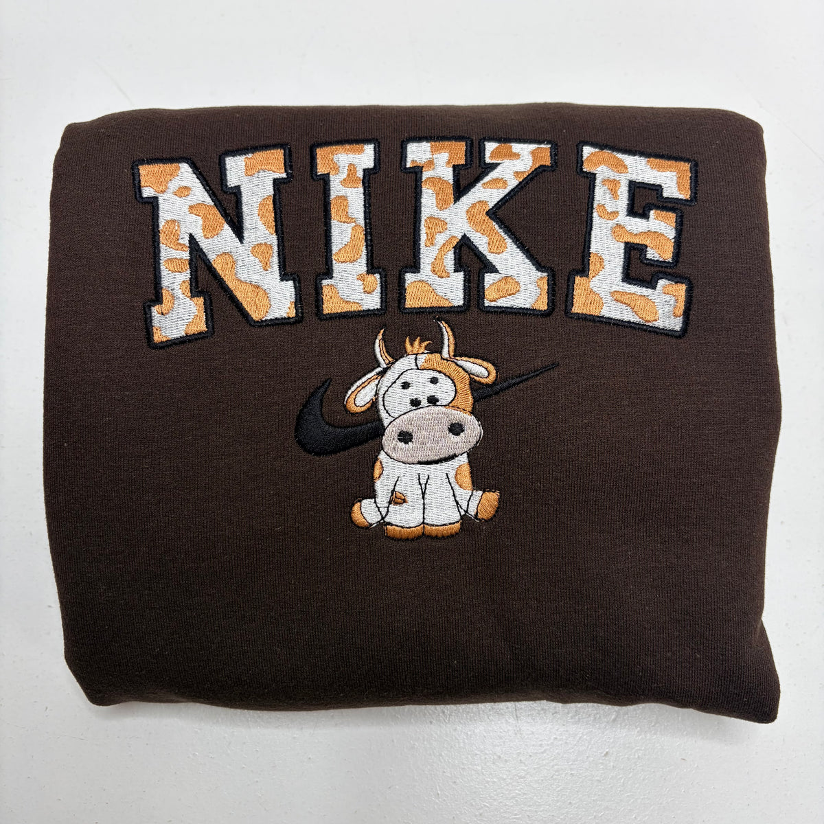 A folded dark brown sweatshirt featuring a large embroidered 'NIKE' logo in orange and white cow-print lettering, with a cartoon cow wearing a black Nike Swoosh underneath.