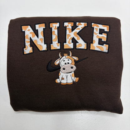 A folded dark brown sweatshirt featuring a large embroidered 'NIKE' logo in orange and white cow-print lettering, with a cartoon cow wearing a black Nike Swoosh underneath.