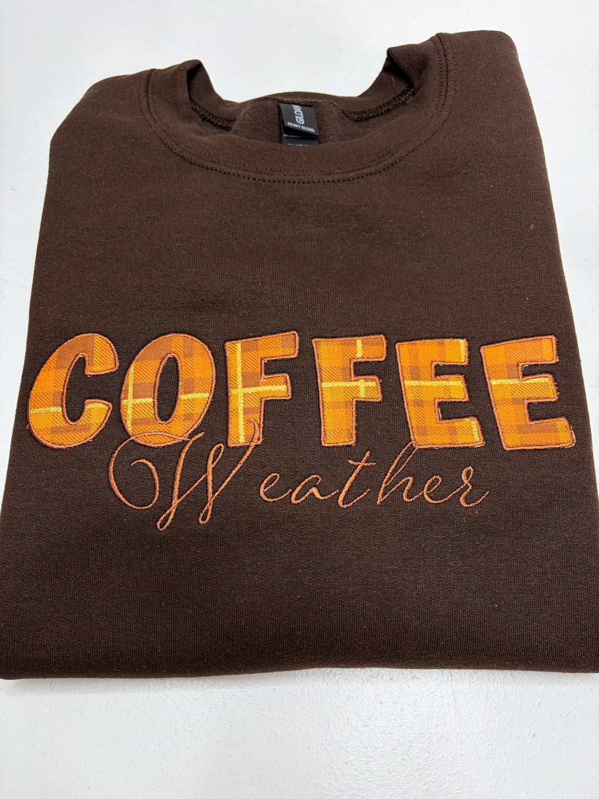 A folded brown crewneck sweatshirt featuring the text 'COFFEE Weather' in orange and yellow plaid and cursive lettering on the front, displayed on a white background.