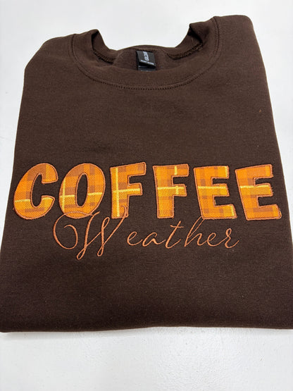A folded brown crewneck sweatshirt featuring the text 'COFFEE Weather' in orange and yellow plaid and cursive lettering on the front, displayed on a white background.