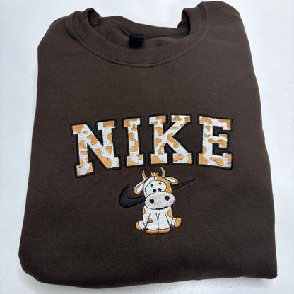 A folded brown crewneck sweatshirt featuring a 'NIKE' graphic with a cow print pattern, a cartoon cow, and a black swoosh on the front, set against a plain white background.
