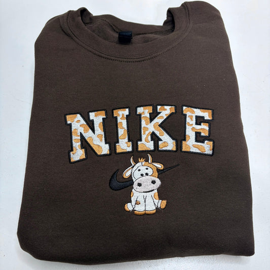 A folded brown crewneck sweatshirt featuring a 'NIKE' graphic with a cow print pattern, a cartoon cow, and a black swoosh on the front, set against a plain white background.