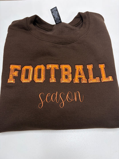 A folded brown crewneck sweatshirt featuring 'FOOTBALL season' embroidered on the front in orange and brown, displayed on a white background.