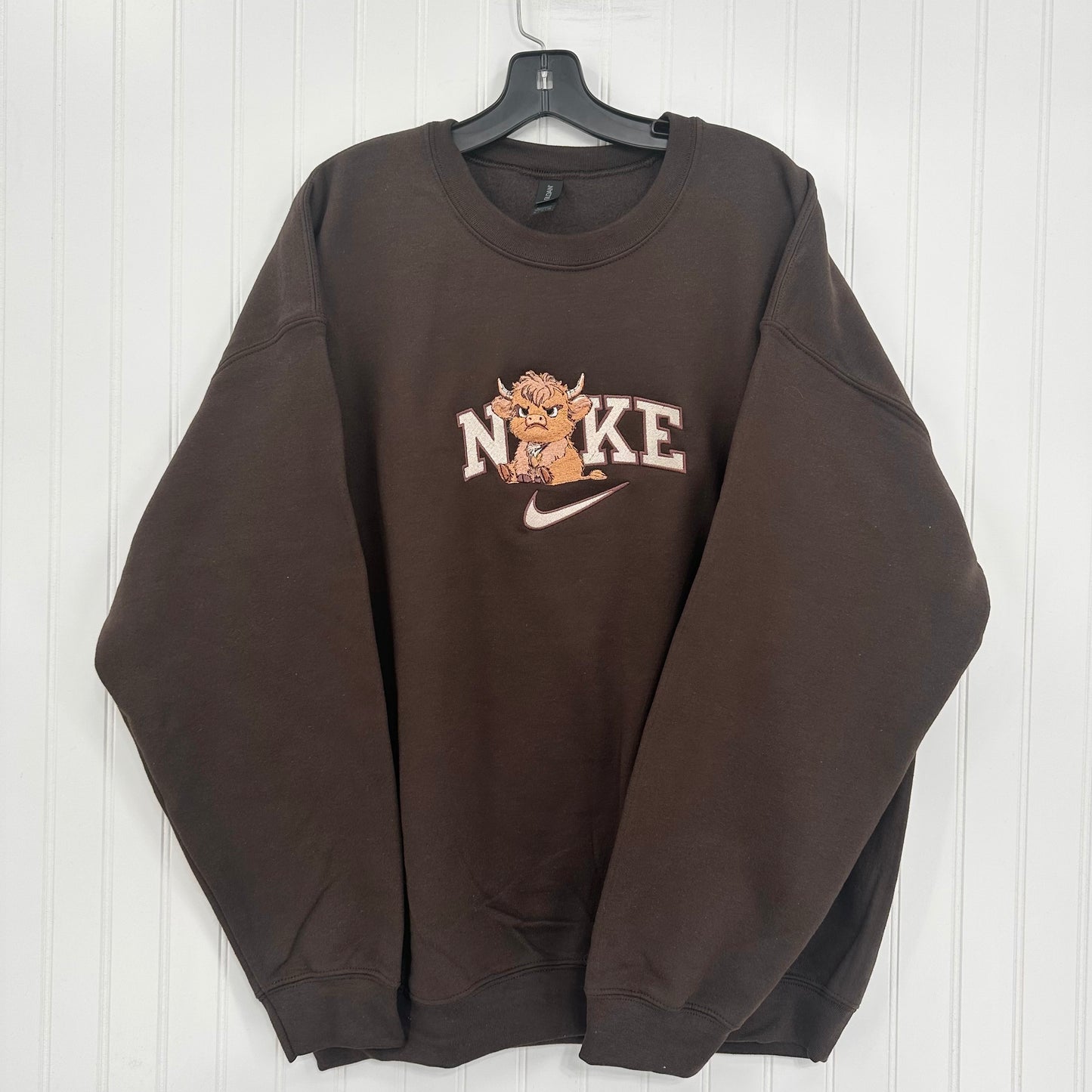 A dark brown crewneck sweatshirt hanging on a black hanger against a white paneled wall. The sweatshirt features a 'NO KE' graphic with a cartoon highland cow and a white Nike swoosh on the chest.