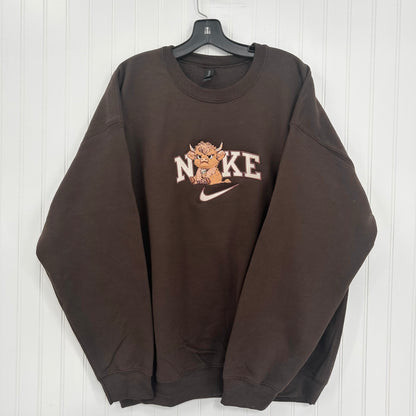 A dark brown crewneck sweatshirt hanging on a black hanger against a white paneled wall. The sweatshirt features a 'NO KE' graphic with a cartoon highland cow and a white Nike swoosh on the chest.