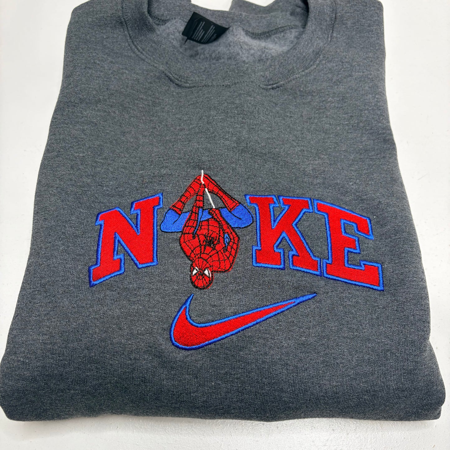 A folded dark gray heather sweatshirt featuring a custom 'N'KE' graphic with a hanging Spider-Man figure and a red and blue Nike swoosh.