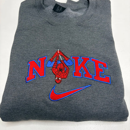 A folded dark gray heather sweatshirt featuring a custom 'N'KE' graphic with a hanging Spider-Man figure and a red and blue Nike swoosh.