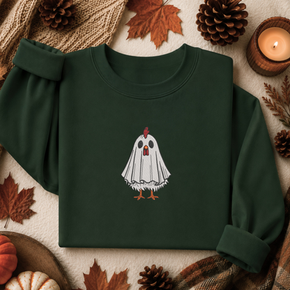 A dark forest green long-sleeve crewneck sweatshirt with a graphic of a white ghost chicken on the chest, styled in a flat lay with autumn props including pinecones, maple leaves, a lit candle, and a knit blanket.