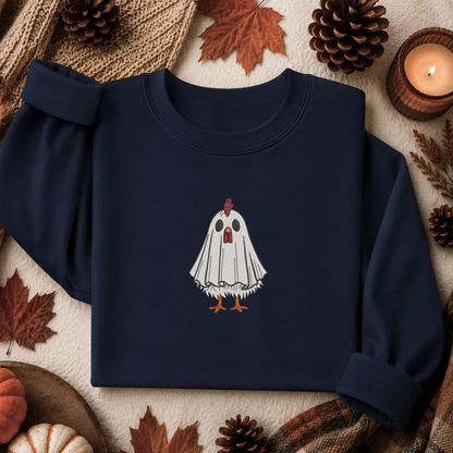 A navy blue crewneck sweatshirt featuring a graphic of a white ghost with orange details, styled in a flat lay with autumn props like pinecones, leaves, and a lit candle.