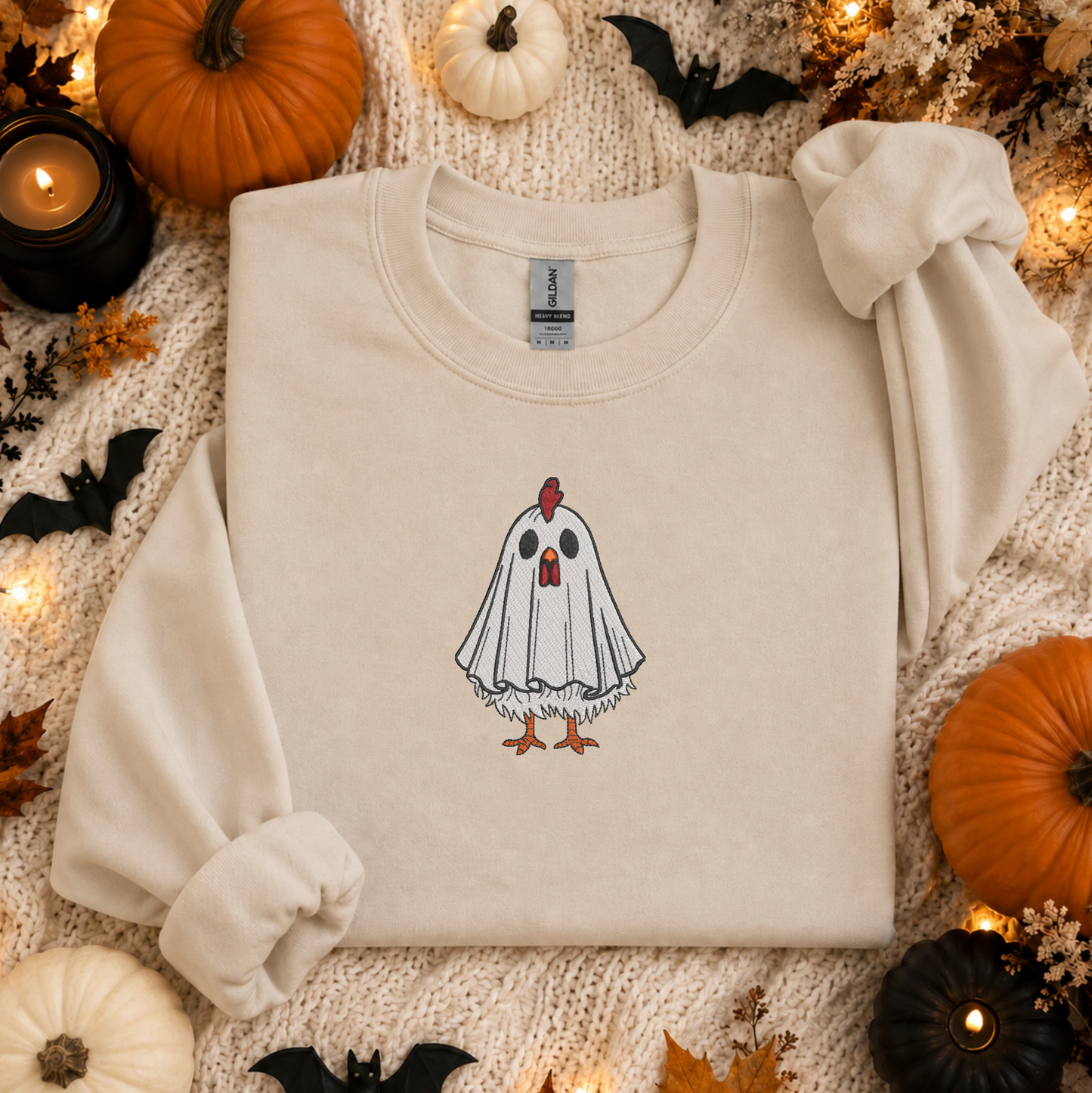 A cream-colored crewneck sweatshirt with a graphic of a ghostly chicken, styled in a cozy autumn flat-lay with pumpkins, candles, and a knitted blanket.