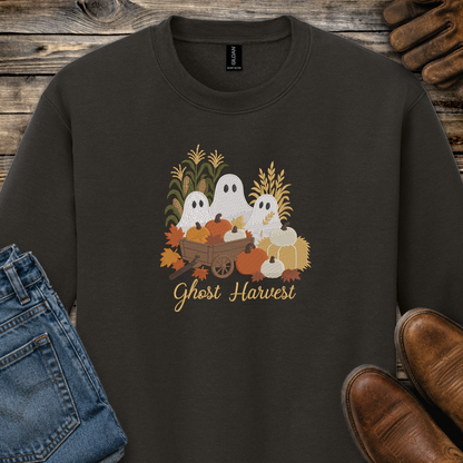 A dark brown crewneck sweatshirt featuring a 'Ghost Harvest' graphic with white ghosts, pumpkins, corn, and autumn leaves. The sweatshirt is styled in a flat-lay arrangement on a rustic wooden background, accompanied by blue jeans, brown leather shoes, and a pair of wooden shoes.