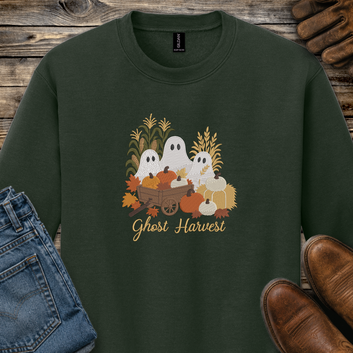 A dark forest green crewneck sweatshirt featuring a 'Ghost Harvest' graphic with cartoon ghosts, pumpkins, corn, and autumn leaves. The sweatshirt is styled in a flat-lay arrangement on a rustic wooden background, accompanied by blue jeans, brown shoes, and a pair of leather gloves.