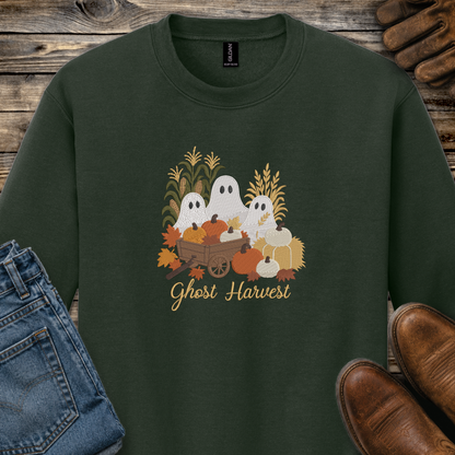 A dark forest green crewneck sweatshirt featuring a 'Ghost Harvest' graphic with cartoon ghosts, pumpkins, corn, and autumn leaves. The sweatshirt is styled in a flat-lay arrangement on a rustic wooden background, accompanied by blue jeans, brown shoes, and a pair of leather gloves.