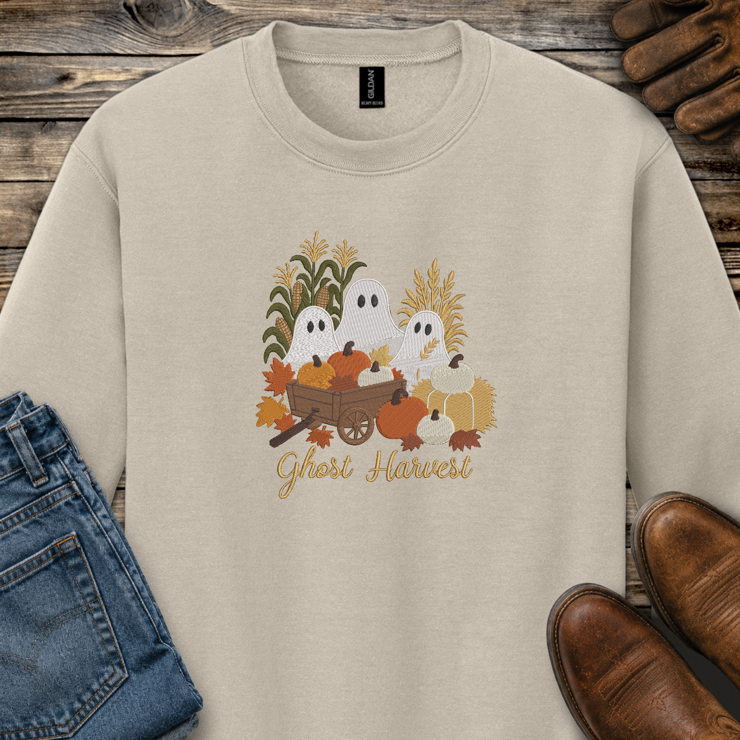 A beige crewneck sweatshirt featuring a 'Ghost Harvest' graphic with cartoon ghosts, pumpkins, corn, and autumn leaves, styled in a flat-lay on a rustic wood background with jeans, shoes, and gloves.