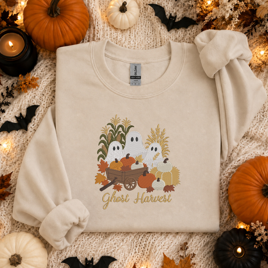 A cream-colored long-sleeve crewneck sweatshirt featuring a 'Ghost Harvest' graphic with cartoon ghosts, pumpkins, corn, and autumn leaves. The sweatshirt is styled in a flat-lay arrangement on a textured cream knit blanket, surrounded by decorative pumpkins, small black bats, and warm string lights.