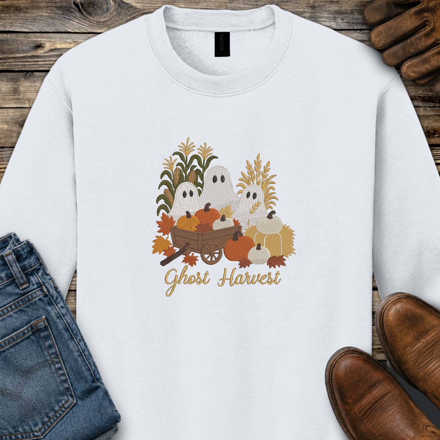 A white long-sleeve crewneck sweatshirt with a 'Ghost Harvest' autumn graphic, styled in a flat-lay on a rustic wooden background with blue jeans and brown leather shoes.