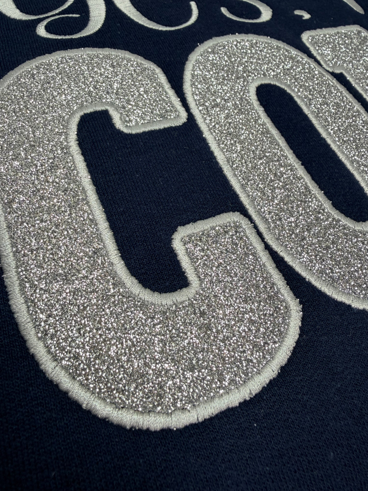 Close-up of a navy blue garment featuring a large glittery silver embroidered letter 'C' with a white stitched outline.