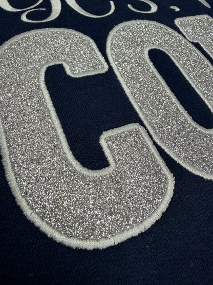 Close-up of a navy blue garment featuring a large glittery silver embroidered letter 'C' with a white stitched outline.