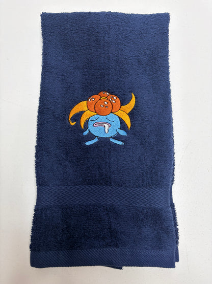 A navy blue terry cloth towel featuring a colorful embroidered pokemon cartoon Gloom character in the center, laid flat against a light off-white background.