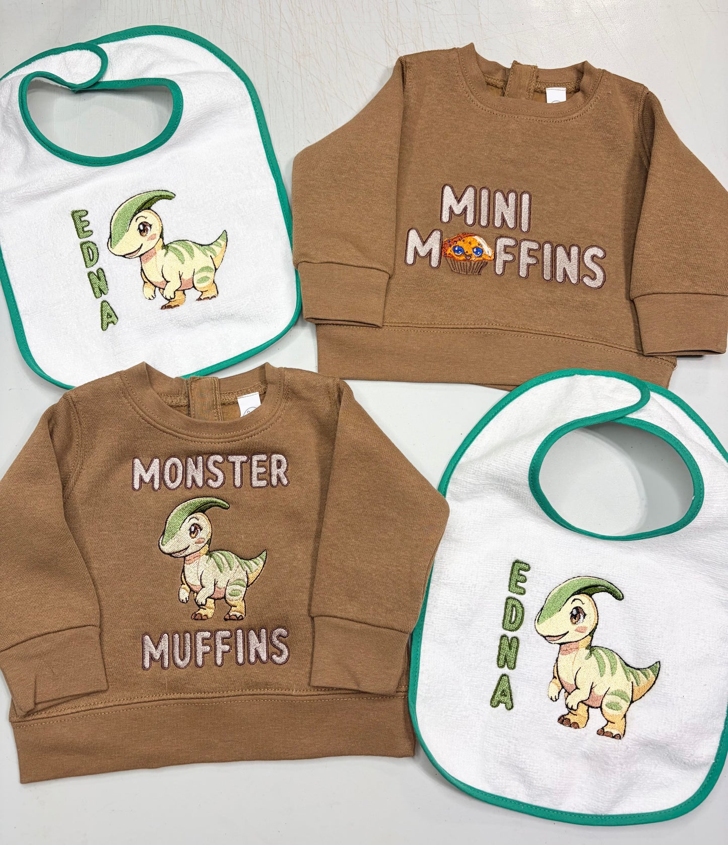 A coordinated baby apparel set laid flat on a light surface, including two brown sweatshirts and two white bibs with green trim. The bibs have a cartoon dinosaur and the text 'EDNA', while the sweatshirts are one brown with 'MINI MuFFINS' and one medium brown with 'MONSTER MUFFINS' and a dinosaur graphic.