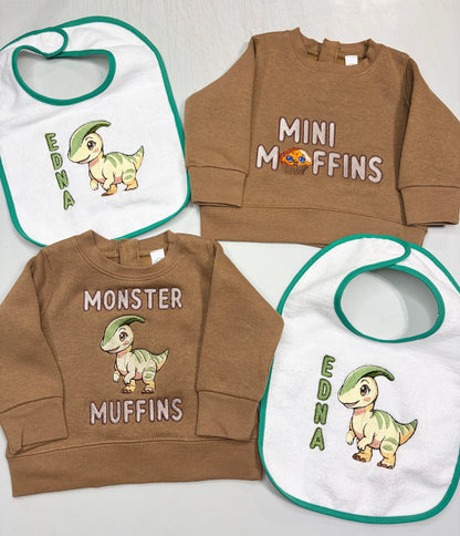 A coordinated baby apparel set laid flat on a light surface, including two brown sweatshirts and two white bibs with green trim. The bibs have a cartoon dinosaur and the text 'EDNA', while the sweatshirts are one brown with 'MINI MuFFINS' and one medium brown with 'MONSTER MUFFINS' and a dinosaur graphic.