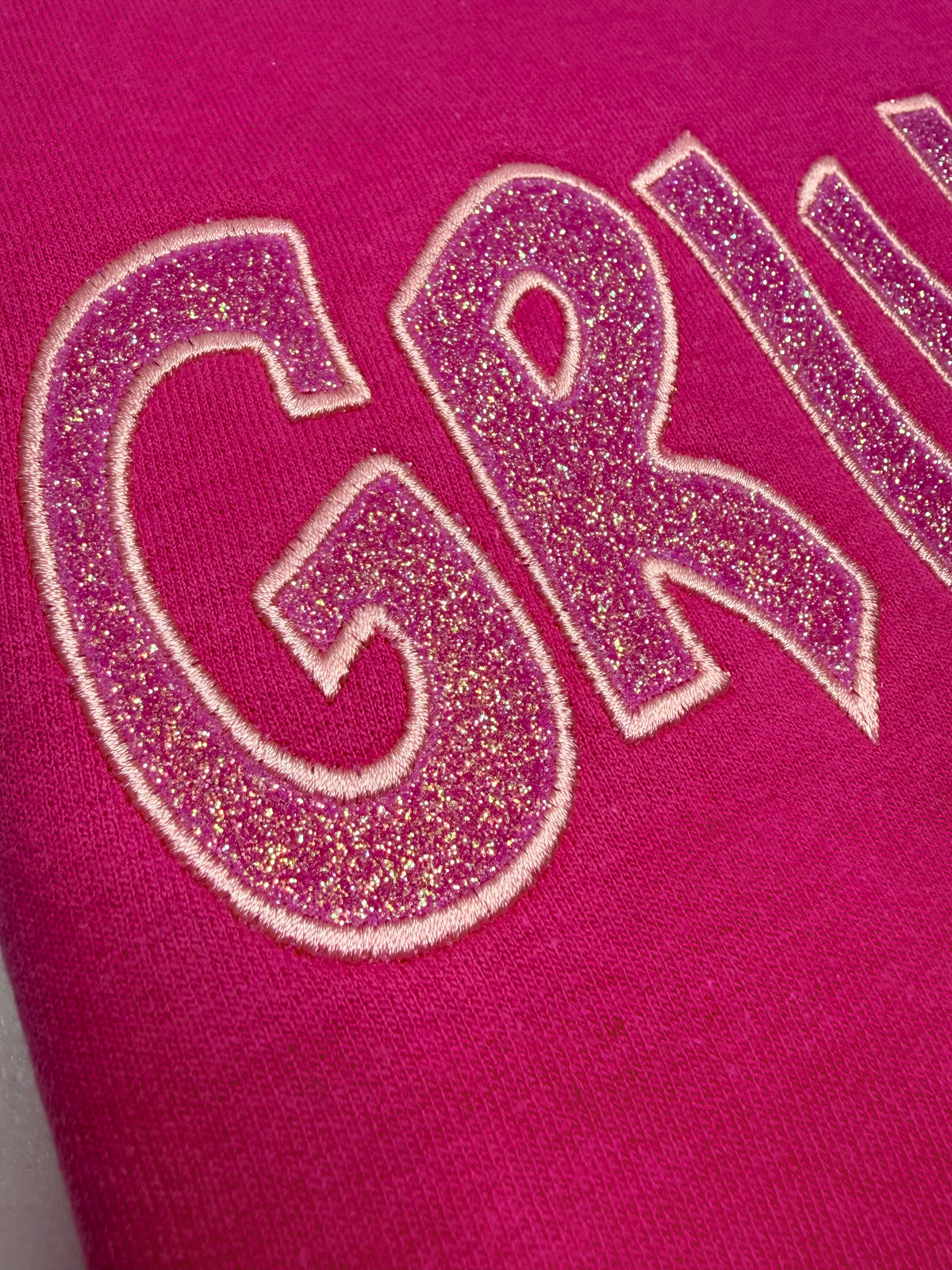 Close-up of a bright pink fabric surface with large glittery gold letters outlined in white, partially showing the word 'GRILL'.